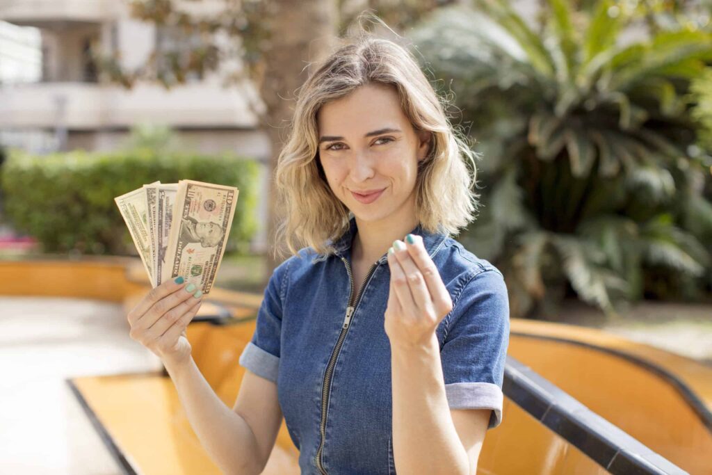 A woman in a denim outfit holds several U.S. dollar bills and gestures with her other hand, outdoors near greenery.