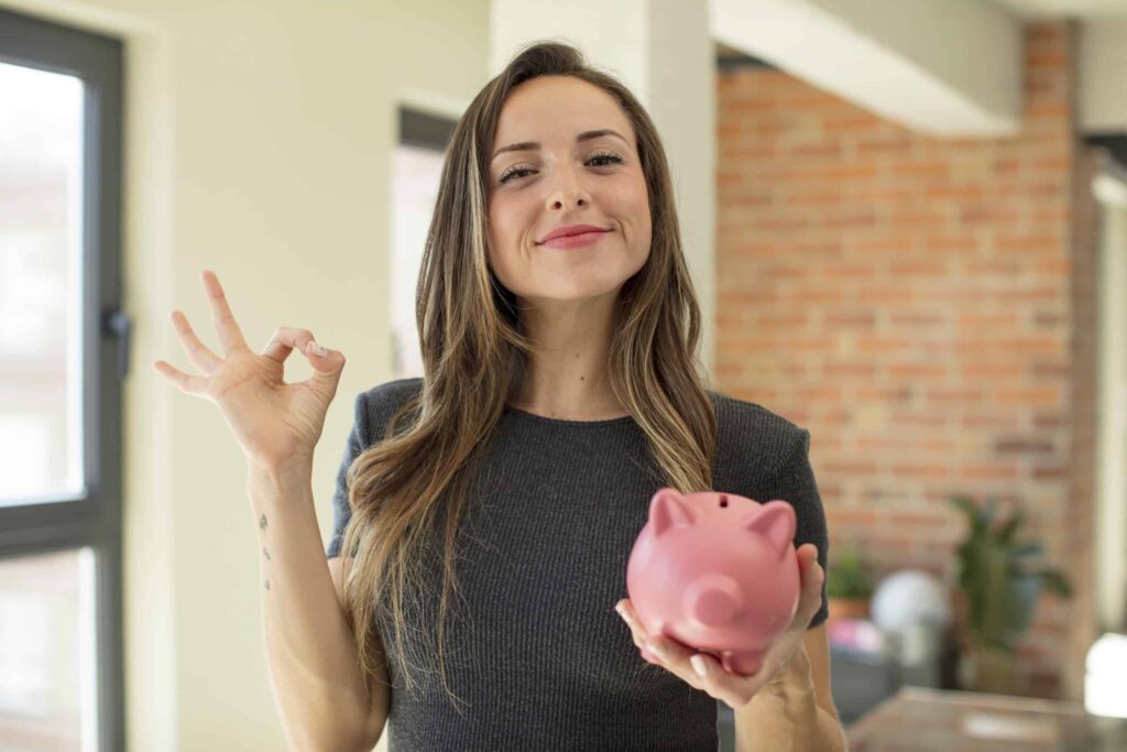 A woman smiles while holding a pink piggy bank in one hand and making an "OK" gesture with the other.