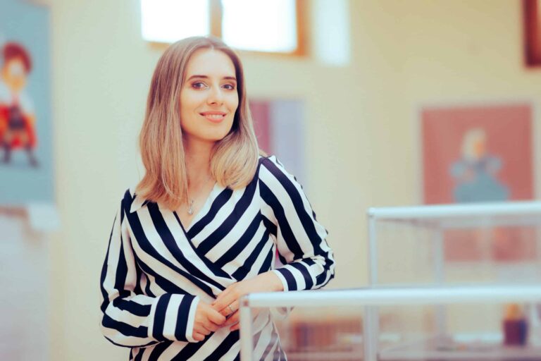 A woman with long hair, wearing a black and white striped top, stands indoors near display cases, smiling at the camera.