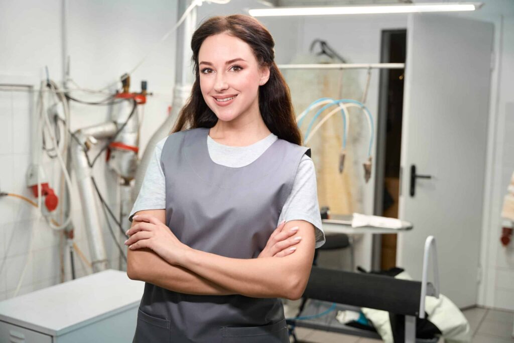 A woman in a work apron smiles while standing with arms crossed in a workshop. Tools and equipment are visible in the background.