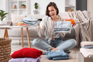Woman sitting on the floor holding folded clothes, surrounded by more folded clothing, in a living room setting.