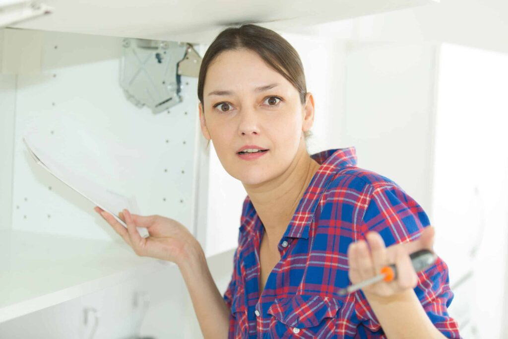 A woman in a plaid shirt holds a paper and screwdriver, looking surprised while standing under an open cabinet.