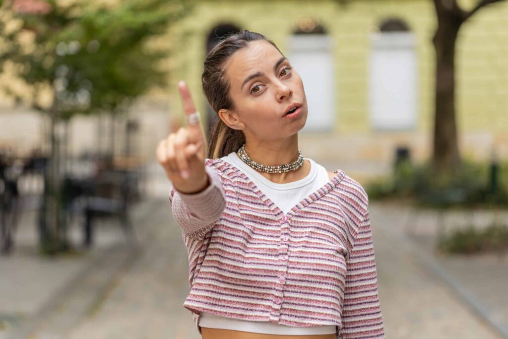 Person outdoors, wearing a striped shirt, pointing with their index finger. Background features a tree and building.