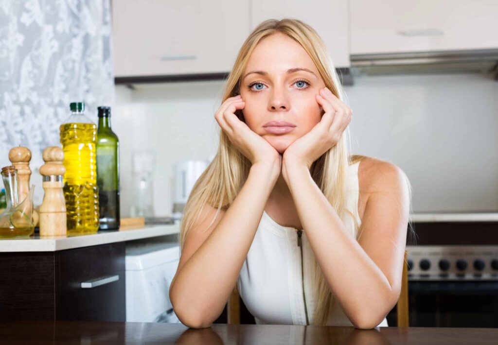 A woman with long blonde hair rests her chin on her hands, sitting at a kitchen table with various bottles and shakers visible in the background.