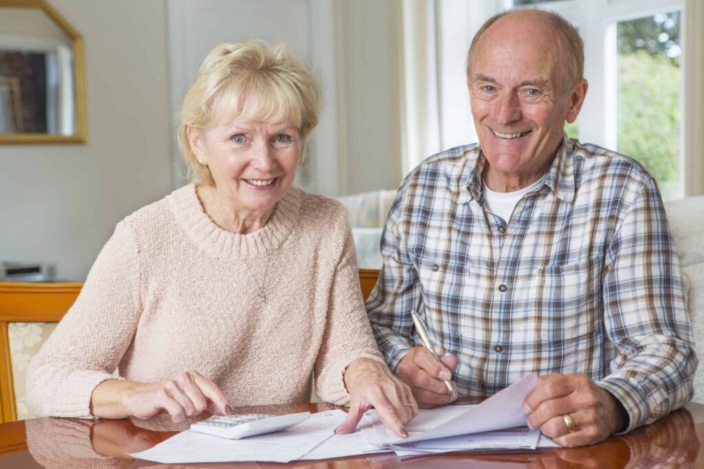 An elderly couple sits at a table working on paperwork, with one holding a calculator and the other a pen.