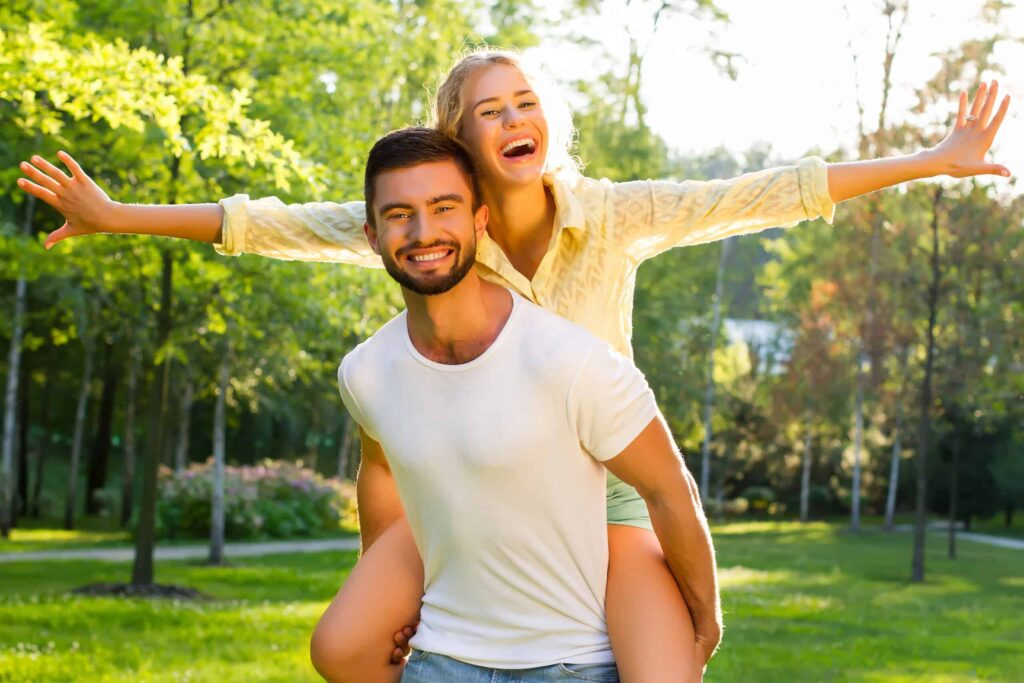 A man gives a woman a piggyback ride in a sunny park. She extends her arms joyfully, both smiling amid green trees and grass.