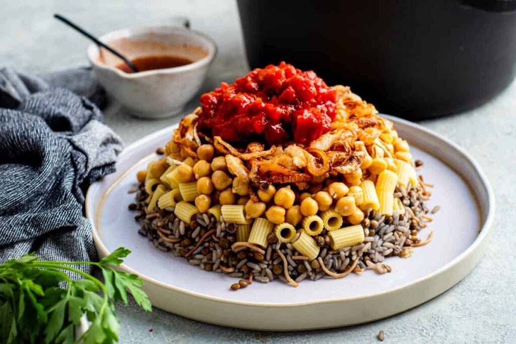A plate of koshari piled with rice, pasta, lentils, chickpeas, crispy onions, and topped with red tomato sauce. A side of sauce and a dark pot are in the background.