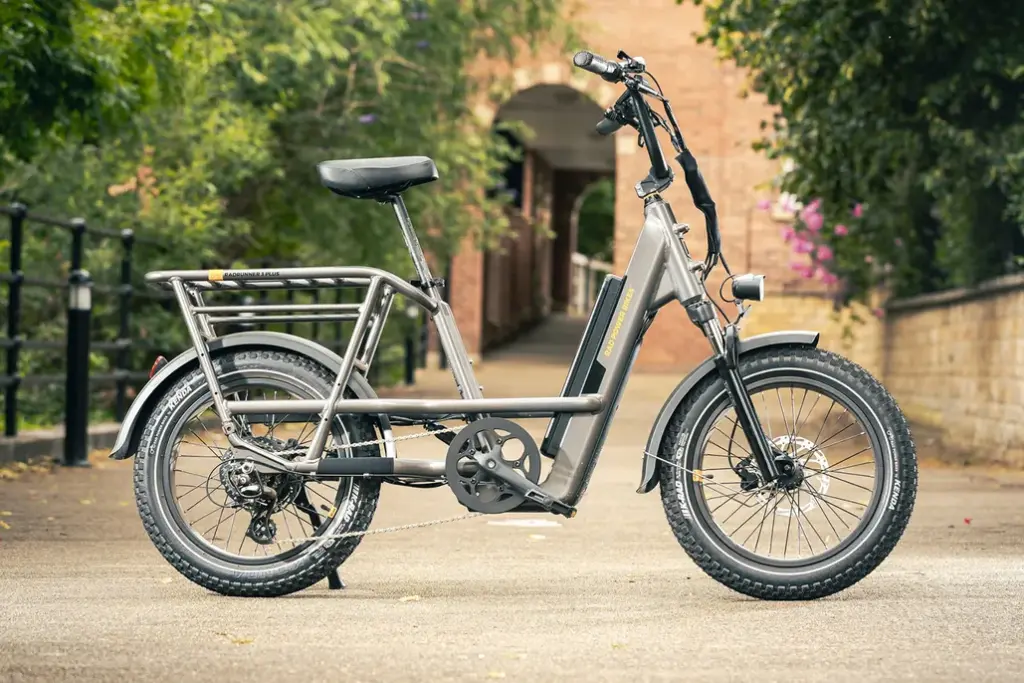 A gray electric bicycle with a sturdy frame, wide tires, and a rear cargo rack is parked on a paved path. A brick archway and greenery are visible in the background.