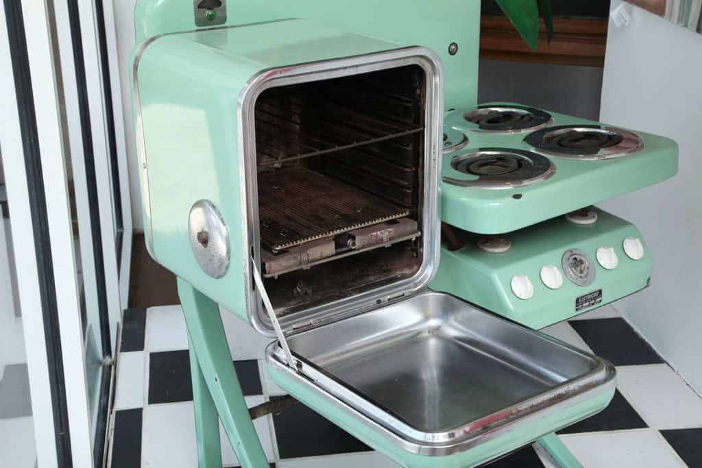 A vintage green stovetop oven with black burners and open oven door on a black and white checkered floor.