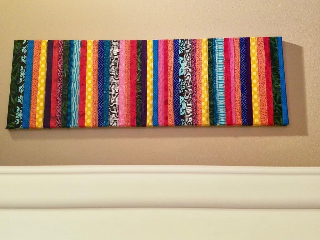 A rectangular textile artwork featuring vertical stripes in various colors and patterns is displayed on a beige wall above a white surface.