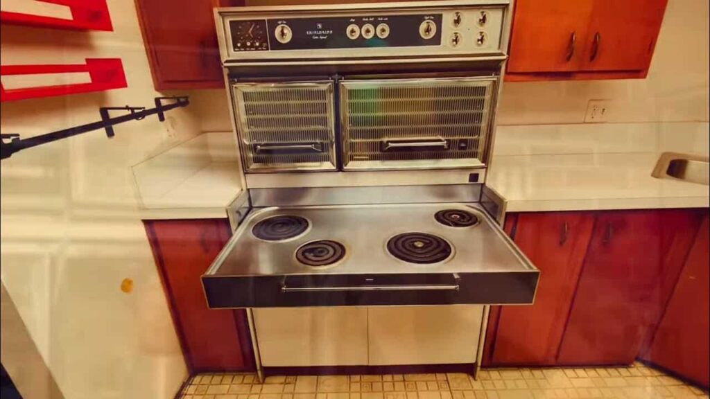 A vintage kitchen with a dual-level electric stove, featuring four coil burners and double ovens. It is surrounded by wooden cabinets and tiled flooring.