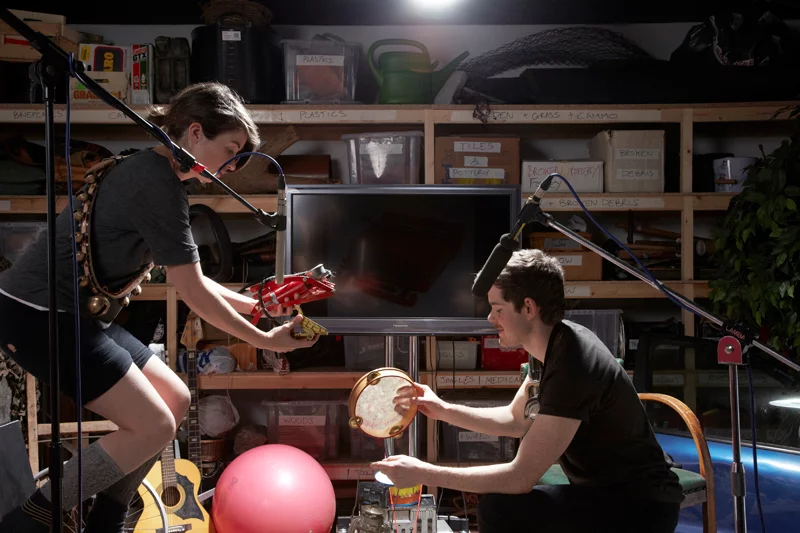 Two people engage in a recording session using various objects, including a guitar, a red toy, and a tambourine, against a backdrop of shelves filled with organized boxes and equipment.
