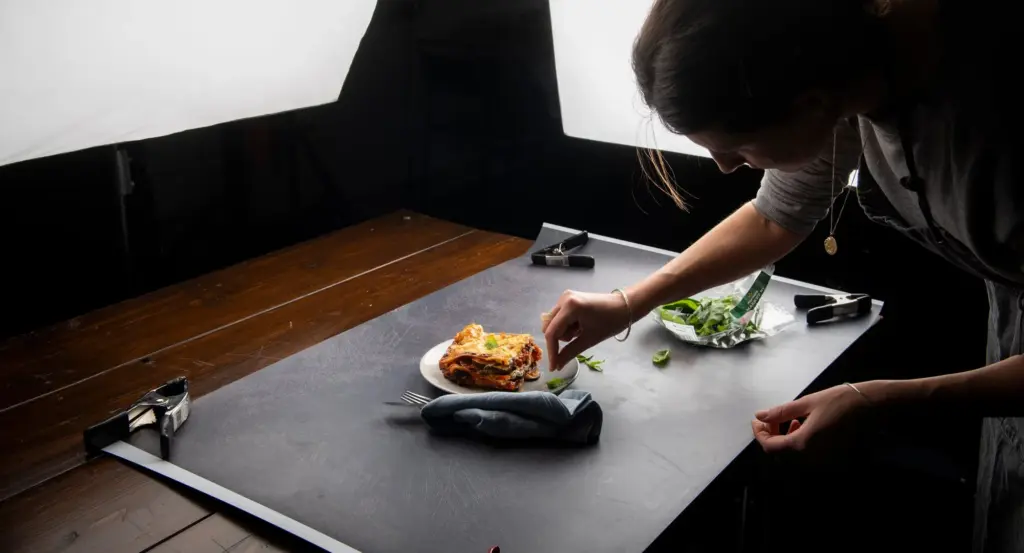 Person arranging a food item on a black surface with bright lights illuminating the scene. A small dish, cloth, and utensils are nearby.