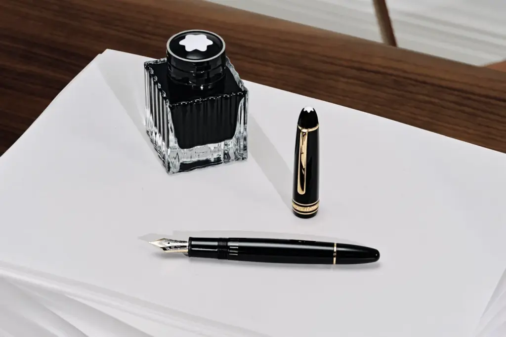 A black fountain pen, an open cap, a square ink bottle, and a stack of white paper on a wooden desk.