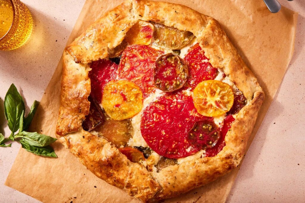 A rustic tomato galette on parchment paper, with a flaky, golden crust, layered slices of red, yellow, and green tomatoes, and fresh basil nearby.