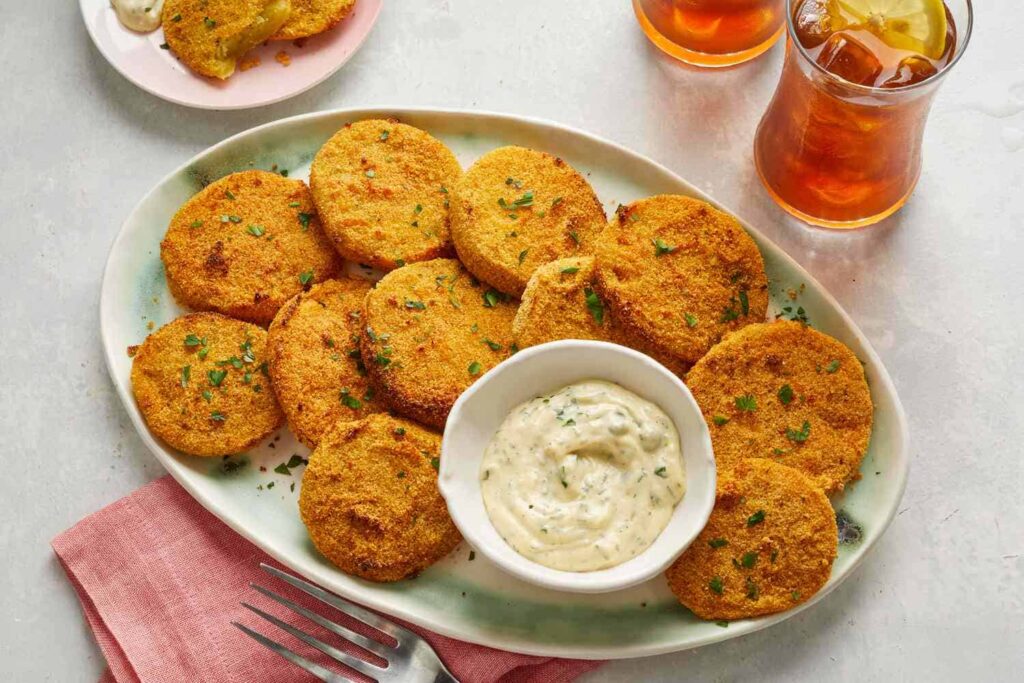 A plate of crispy fried green tomatoes with a dipping sauce in the center, garnished with herbs. Two glasses of iced tea are in the background.