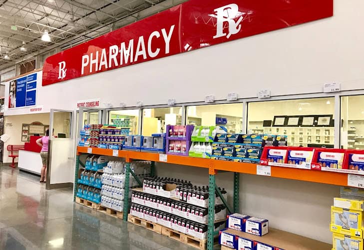 Pharmacy section in a store with shelves displaying various medical supplies and products. People are seen at the counter in the background.