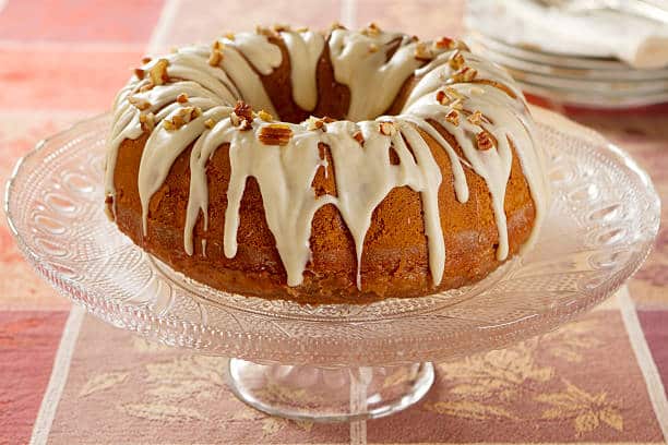 Bundt cake with white icing and chopped nuts on a glass cake stand.