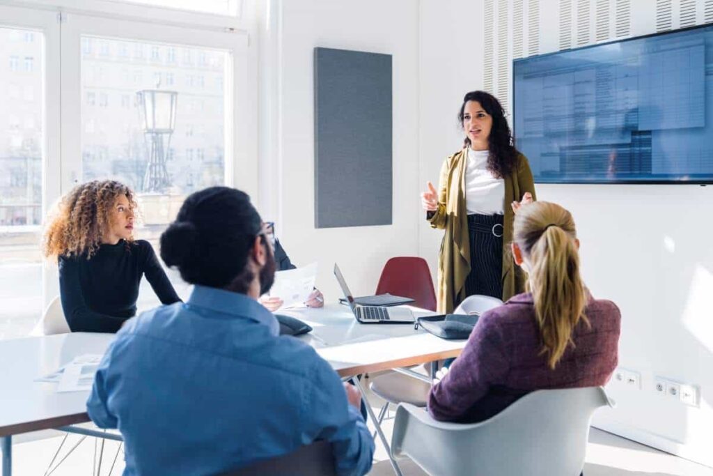A woman stands and speaks to four seated colleagues during a meeting in a bright conference room with a large screen on the wall.