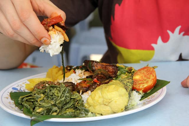 A person eating nasi campur, a plate of rice with various side dishes including vegetables, omelet, and shrimp.