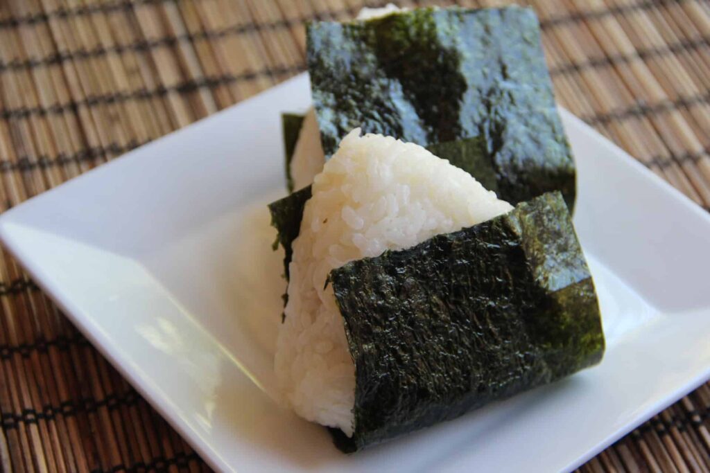 Two triangular rice balls wrapped in nori seaweed are placed on a white square plate, set against a bamboo mat background.