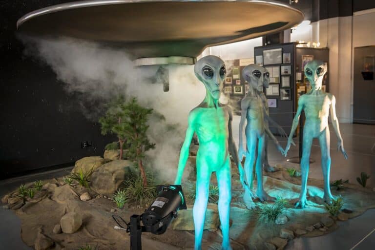 Three alien figures stand under a UFO model with smoke effects in a museum display.