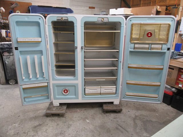 Open vintage refrigerator with multiple compartments and shelves in a workshop setting.