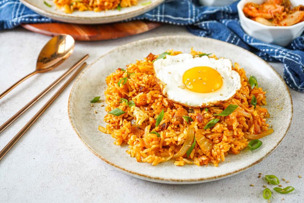 A plate of fried rice topped with a sunny-side-up egg, garnished with green onions. A small bowl of side dish is in the background, next to a fork and a spoon.
