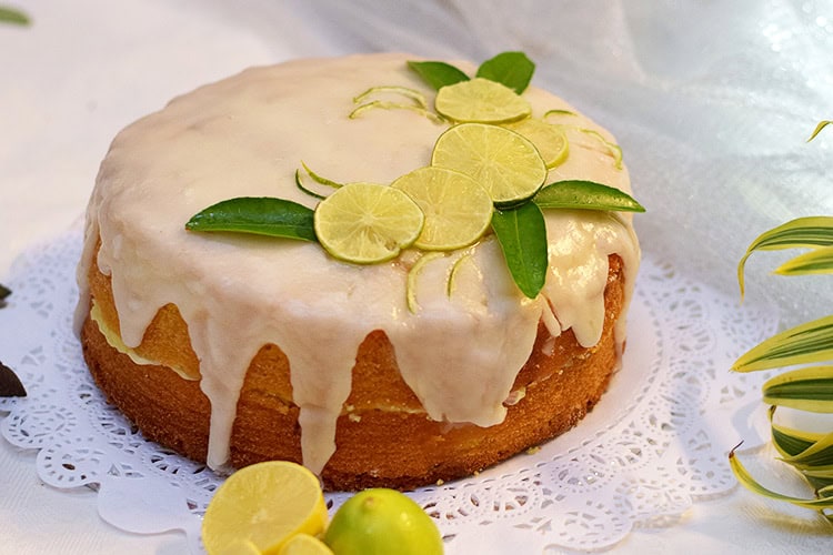 Lemon cake topped with glaze, lime slices, and green leaves on a white doily.