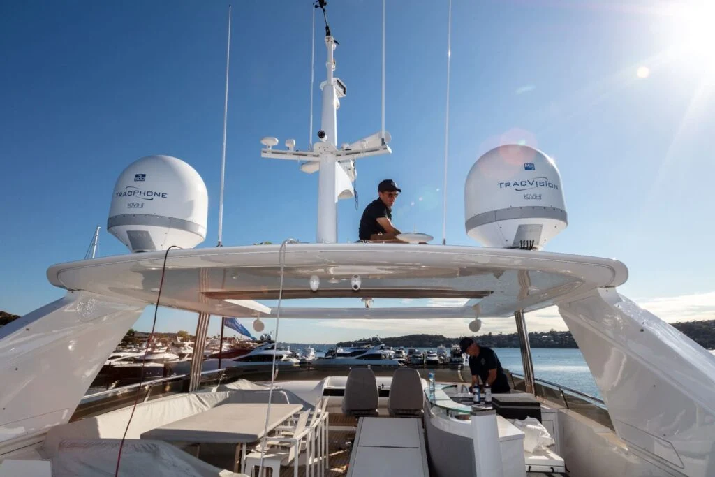 Two people work on a yacht's upper deck under clear skies. The deck features satellite equipment and seating. Boats and hills are visible in the background.