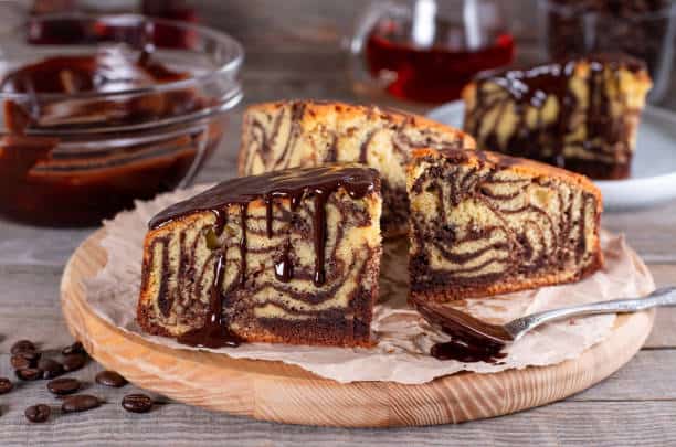 A sliced marble cake with chocolate glaze on a wooden board, surrounded by bowls of chocolate and a fork.