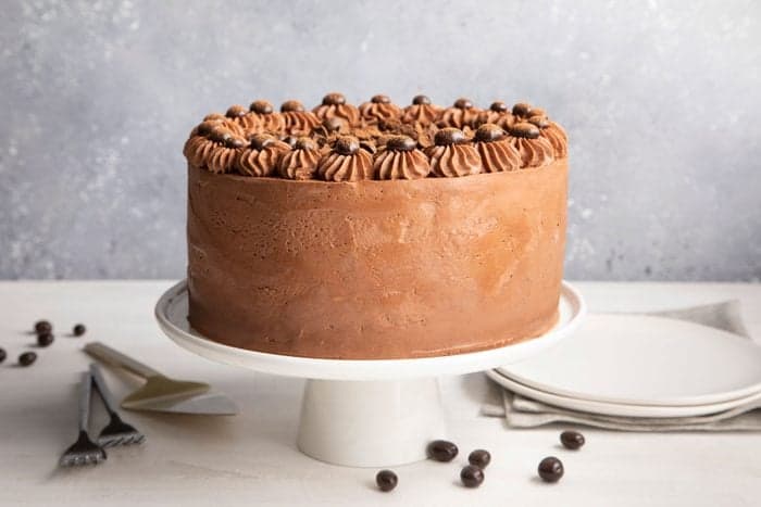 Chocolate cake with frosting and decorative chocolate chips on top, placed on a white cake stand with utensils and plates nearby.