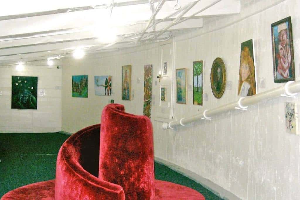 Art gallery with white walls displaying various paintings. A central spiral red velvet structure is in the foreground.