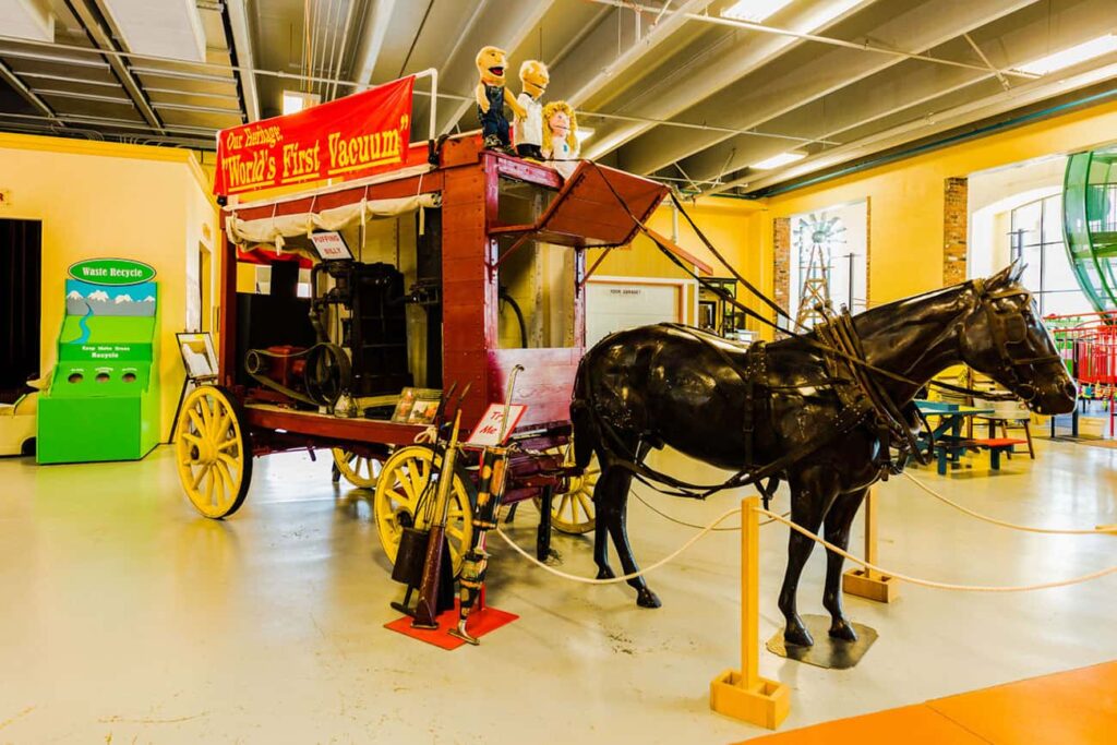 A vintage horse-drawn vacuum cleaner carriage displayed indoors with informational signs and figures on top.