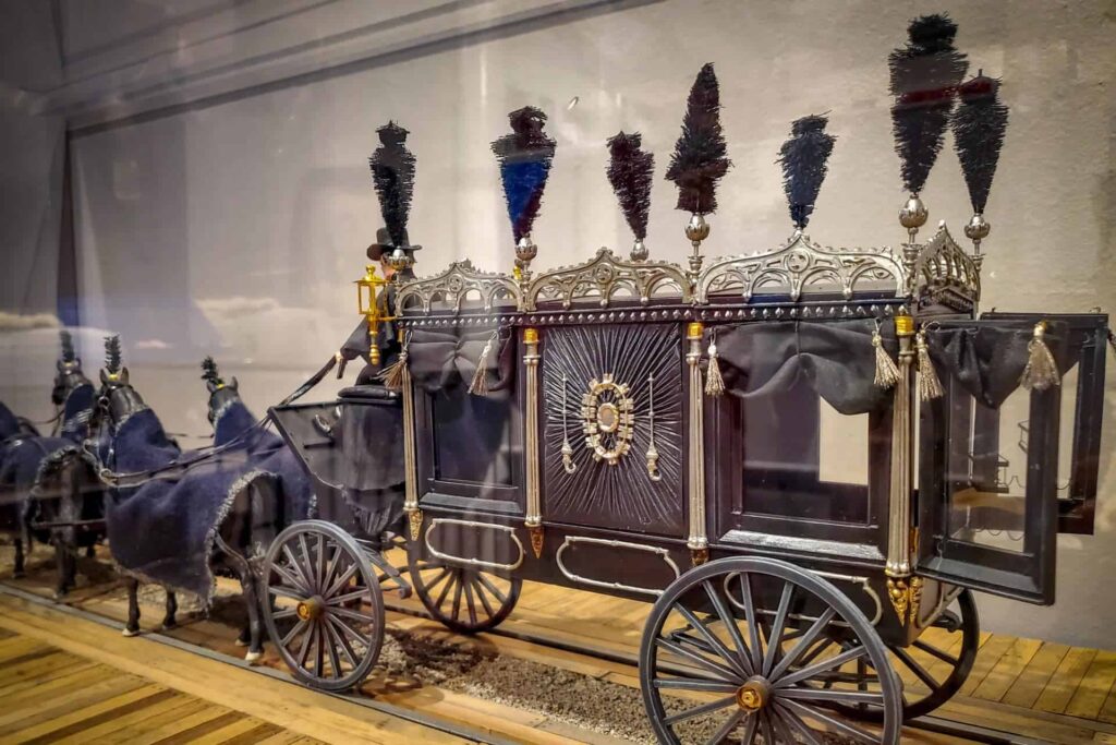 An ornate, vintage horse-drawn funeral carriage with decorative plumes and black drapery is displayed indoors.
