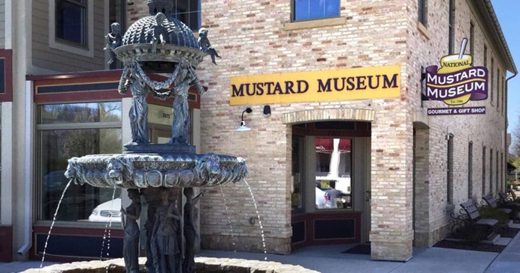 A brick building with a sign reading "Mustard Museum" features a decorative stone fountain in front, with water flowing.