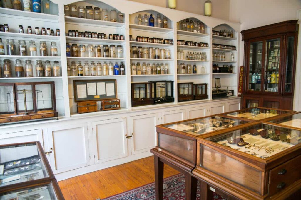 A vintage pharmacy interior with shelves displaying various bottles and jars, and glass display cases containing historical medical instruments.