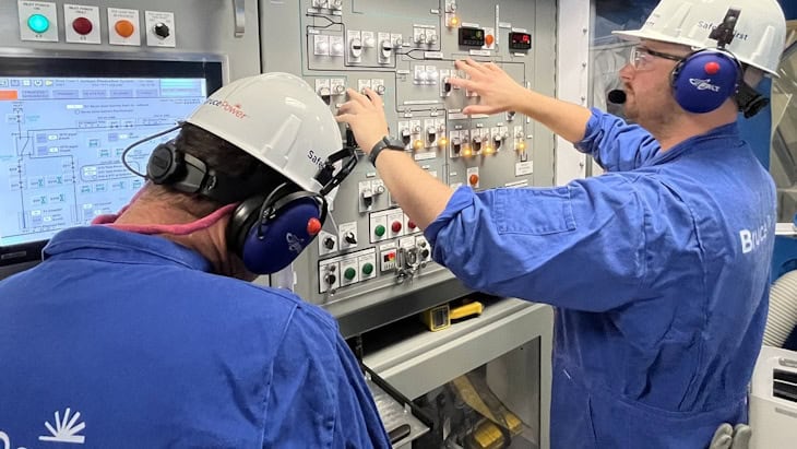 Two workers in blue uniforms and helmets operate a control panel with switches and buttons, wearing headphones in an industrial setting.