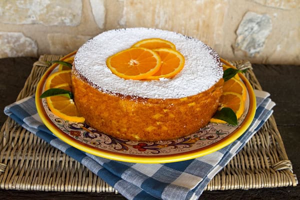 Round cake topped with powdered sugar and orange slices on a decorative plate over a blue checkered cloth.