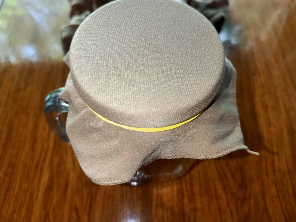A glass jar covered with a brown cloth, secured by a yellow elastic band, sits on a wooden surface.