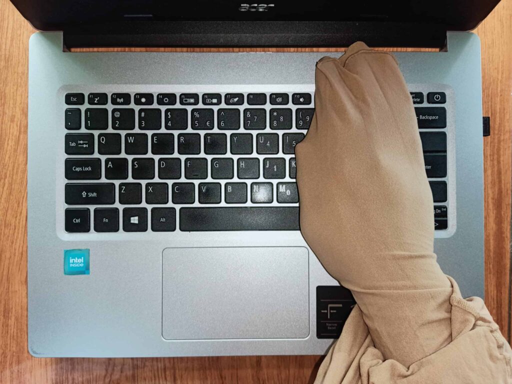 A gloved hand types on a silver laptop keyboard with a wooden surface background.