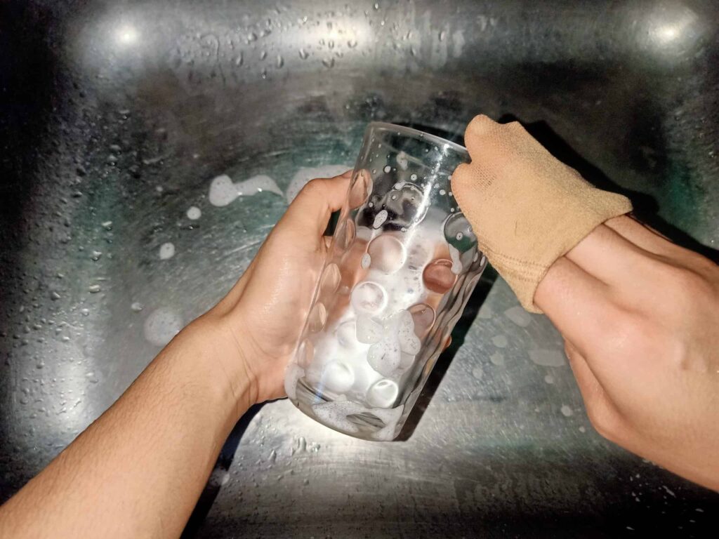 Two hands washing a clear glass in a stainless steel sink with soap suds.