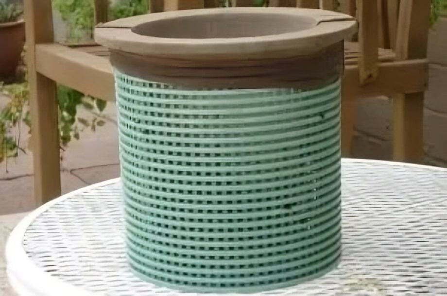 A green and brown striped clay pot sits on a white metal table outdoors, with a wooden chair and potted plants in the background.