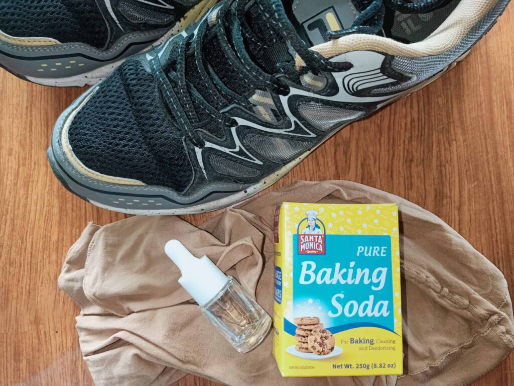 A pair of athletic shoes, a box of baking soda, a small dropper bottle, and a piece of beige fabric are placed on a wooden surface.