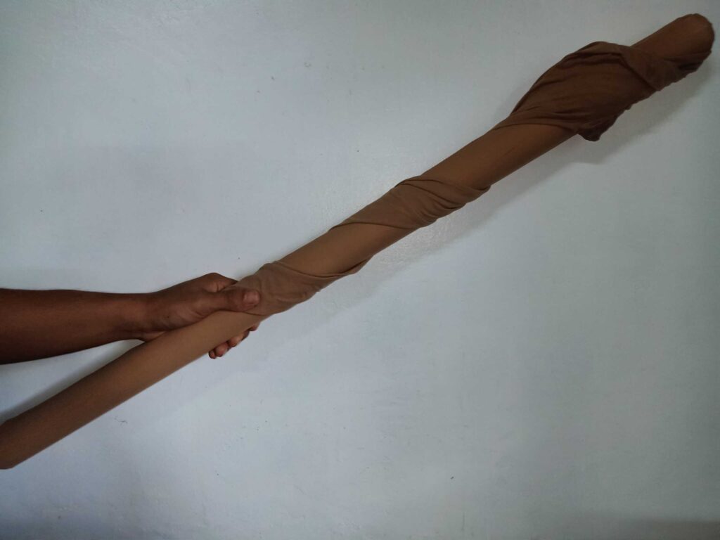 A hand holds a long cylindrical object wrapped in brown fabric against a plain wall.