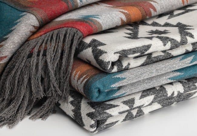 Stack of patterned woven textiles in various colors, including blue, orange, and gray, with fringe on one end.