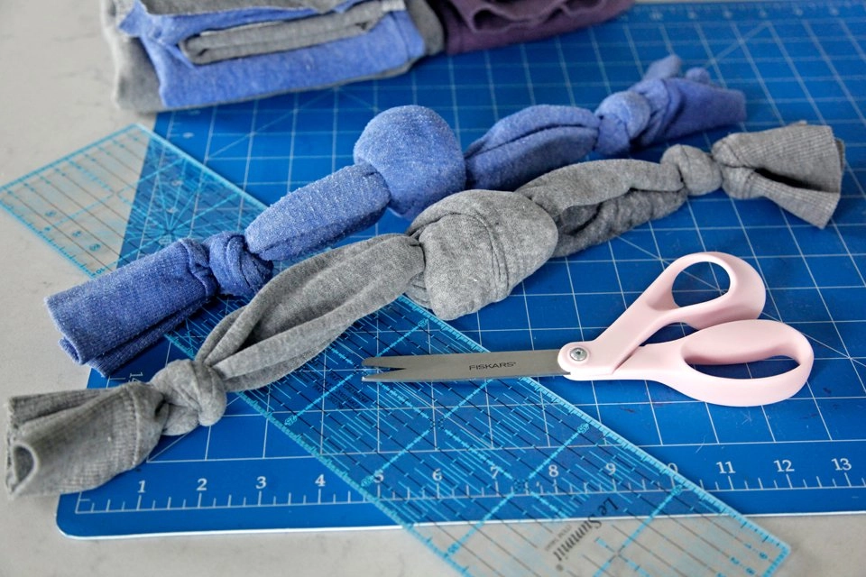 Two braided fabric knots, one blue and one gray, laid on a blue cutting mat with rulers and pink scissors.