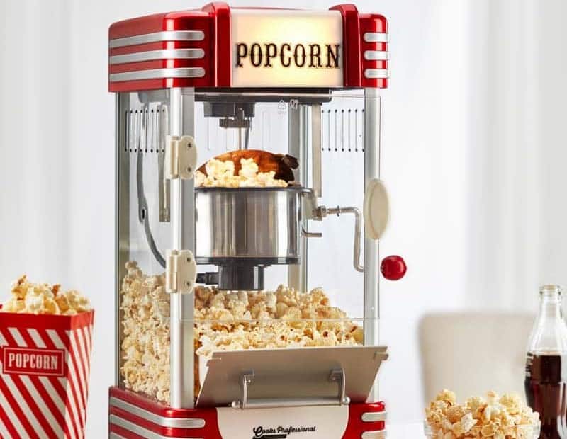 A red popcorn maker with a vintage design, filled with popcorn. A striped popcorn box and a bottle are visible beside it.