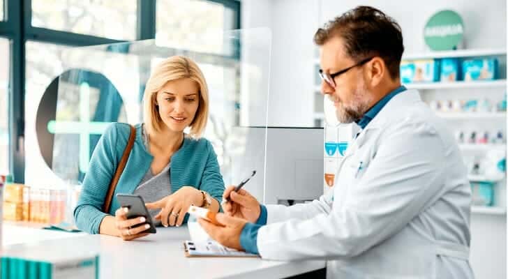 A woman at a pharmacy counter speaks with a pharmacist. She is holding a smartphone, while the pharmacist in a white coat examines a prescription.