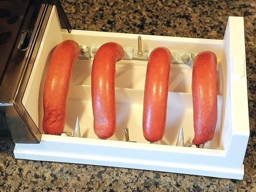 Four hot dogs placed on metal skewers inside a white kitchen appliance on a granite countertop.